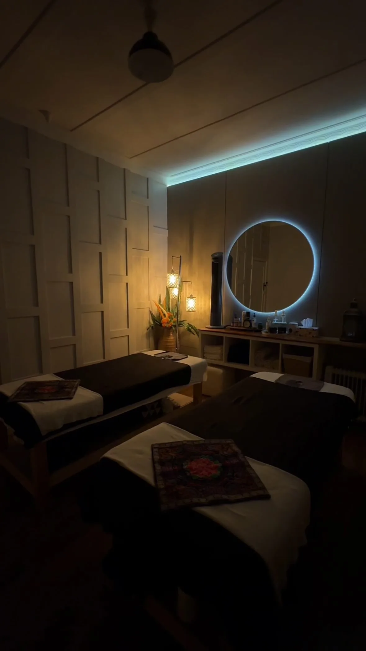 A Siam Ivory treatment room — two tables dressed in Thai runners, backlit round mirror, warm floor lantern